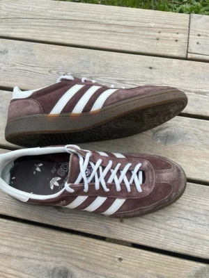 Adidas Originals bruna sneakers - Snygga Adidas Originals sneakers i mocka med brun färg och klassiska vita ränder på sidorna. Skorna har vit hälkappa, vita skosnören och en gummisula med retrovibbar. Perfekta för dig som gillar streetstyle och tidlös design.