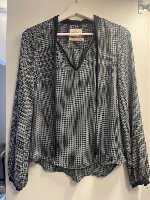 Mönstrad blus från Holly & Whyte  - Snygg svartvit blus från Holly & Whyte  med zigzag-mönster, v-ringning och dekorativa band vid halsen. Långärmad modell med mjukt, lätt tyg och svarta detaljer vid ärmslut och krage. Perfekt för en trendig och avslappnad look.