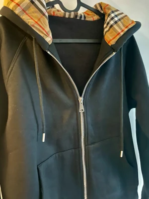 Svart hoodie med Burberry-inspirerad huva - Svart hoodie med dragkedja och fickor framtill. Huvan har ett klassiskt rutigt foder i beige, svart, vitt och rött som påminner om Burberrys ikoniska mönster. Tillverkad i mjukt bomullsmaterial och har snören i huvan för justering. Perfekt för en chill och stilren look.