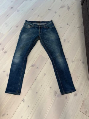 Blå raka jeans från Nudie Jeans - Snygga blå jeans från Nudie Jeans med raka ben och klassisk femficksdesign. Jeansen har slitningar och ljusare partier framtill och bakfickor med dekorativa sömmar. Perfekta för en avslappnad och trendig look.