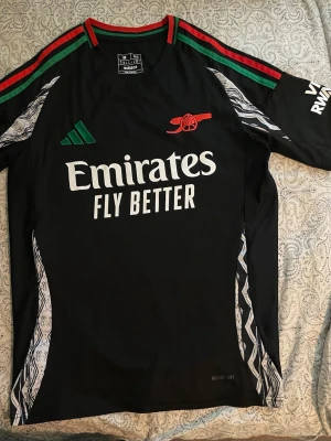 Arsenal svart tränings t-shirt Adidas - Säljer en original Arsenal-tröja som jag köpte i vintras i samband med ett besök på Emirates Stadium i London. Tröjan är köpt direkt i klubbens officiella butik vid arenan.  Tröjan är i storlek M och är i mycket fint skick, eftersom den knappt är använd. Säljer den då den tyvärr blivit lite för liten för mig. 