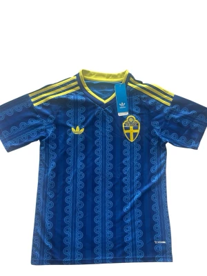 Blå Sverige fotbollströja Adidas S - Blå fotbollströja från Adidas med Sveriges landslagsemblem på bröstet och gula detaljer. Tröjan har korta ärmar, V-ringning med gul kant och tre gula ränder på axlarna. Tillverkad i lätt och ventilerande material, perfekt för träning eller match.