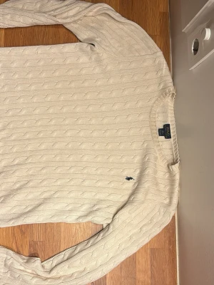 Kabelstickad beige tröja Polo Ralph Lauren - Kabelstickad beige tröja från Polo Ralph Lauren med rund halsringning och lång ärm. Tröjan har det klassiska mörkblå broderade logotypen på bröstet och är tillverkad i mjukt material. Perfekt för lager på lager och stilren look. Tröjan är XL i barn men sitter som S i vuxen. Billigare pris vid snabb affär 