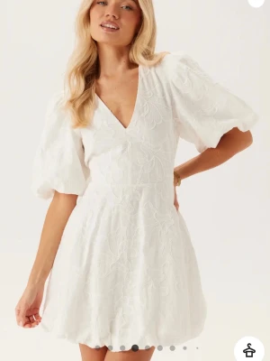 Vit kort klänning med puffärm - Säljer denna superfina slutsålda vita kortklänningen med v-ringning, puffärmar och knyt i ryggen. Nyskick! Aldrig använd med lappar kvar. Perfekt till midsommar eller studenten! 🌸