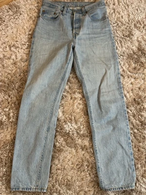Levi's ljusblå raka jeans - Säljer ett par klassiska ljusblå Levi's jeans 501 med rak passform och hög midja. Jeansen har fem fickor, knappgylf samt den ikoniska läderlappen bak i midjan. Tillverkade i slitstark denim av bomull. Byxorna har en liten lagning under ena bakfickan (se bild). Storleken är W26 L30