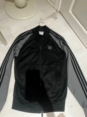 Hoodie adidas - Säljer en svart Adidas track jacket med grå ärmar och klassiska tre ränder längs axlarna. Dragkedja framtill och Adidas logga broderad på bröstet. Perfekt för en sportig och avslappnad stil.