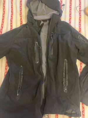 Svart vindjacka från Haglöfs - Snygg svart vindjacka från Haglöfs med huva och flera dragkedjeförsedda fickor framtill och på ärmen. Jackan har en sportig look och är tillverkad i ett slitstarkt material som står emot vind. Perfekt för dig som gillar outdoor och streetwear.