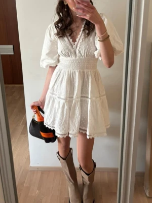Vit kort klänning med puffärm - Söt vit kort klänning med puffärmar, v-ringning och smock i midjan. Klänningen har spetsdetaljer och volangkant nertill samt diskret prickigt mönster. Perfekt för sommarens alla tillfällen.