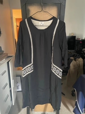 Svart klänning med broderi Noa Noa - Svart klänning från Noa Noa med vita broderade detaljer längs sidorna och på fickorna. Klänningen har trekvartsärm och rund halsringning. Perfekt för dig som gillar en enkel men unik stil.