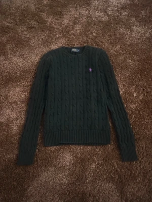 Mörkgrön kabelstickad tröja Polo Ralph Lauren - Mörkgrön kabelstickad tröja från Polo Ralph Lauren med lila broderad logga på bröstet. Tröjan har rund halsringning, långa ärmar och ribbade muddar vid ärmslut och nederkant. Klassisk och tidlös design i mjukt material.
