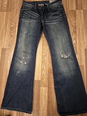 Blå bootcut jeans från crocker - Strl 28/32. Säljer ett par mörkblå bootcut jeans från crocker med slitningar och distressed detaljer på framsidan. Jeansen har klassisk femficksmodell, låg midja och tydliga whiskers vid höfterna. Tillverkade i kraftig denim med vintagekänsla.