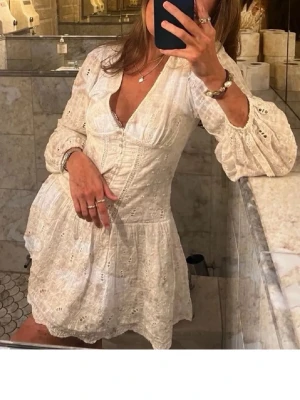 Zara studentklänning - Söt vit kortklänning med v-ringning, spetsdetaljer och knappar framtill. Klänningen har långa puffärmar med resår vid ärmslut och volang nedtill. Perfekt för sommarens eller studenten