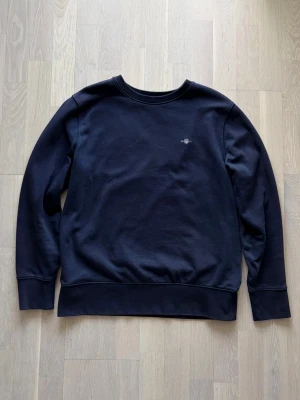 Mörkblå sweatshirt från Gant - Säljer en stilren mörkblå sweatshirt från Gant med rund halsringning och diskret logga på bröstet. Tröjan har långa ärmar och breda muddar vid ärmslut och nederkant. 