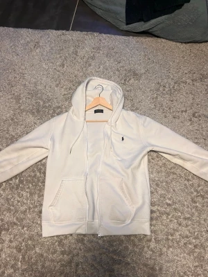 Vit hoodie från Polo Ralph Lauren - Snygg vit hoodie med dragkedja från Polo Ralph Lauren. Klassisk design med huva och dragsnören, två fickor framtill och den ikoniska lilla loggan broderad på bröstet. Perfekt för en clean och avslappnad stil.