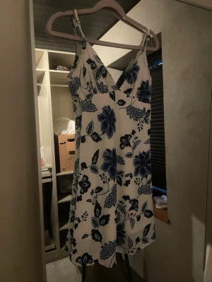 Vit kortklänning med blå blommor - Söt vit kortklänning med blått blommönster och smala axelband. Klänningen har v-ringning och är ärmlös, perfekt för varma dagar. Materialet känns lätt och luftigt, vilket gör den skön att bära. Passformen är normal och modellen är feminin och somrig.