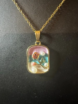 Guldhalsband med ros och pärlor - Unikt halsband med guldfärgad kedja och rektangulärt hänge. Hänget har en inramad design med en liten röd ros, gröna blad och flera vita pärlor mot en bakgrund i rosa, blått och beige. Perfekt för dig som gillar detaljerade och färgglada smycken.