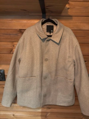 Beige ullrock från 157 - Stilren beige ullrock från 157 med rak passform och stora framfickor. Rocken har klassisk krage, knäppning framtill och quiltat foder på insidan för extra värme. Perfekt för kyliga dagar och enkel att matcha med olika outfits.