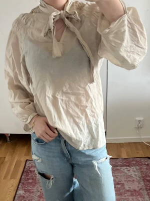 Beige linne blus med knytband i halsen - Säljer en luftig beige linne blus med knytband i halsen och lätt puffade axlar. Blusen har lång ärm och är tillverkad i ett tunt, något transparent tyg som ger en avslappnad och trendig look. Perfekt att styla med jeans eller kjol. Pris kan diskuteras🩷