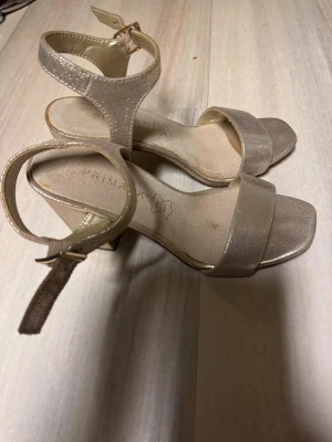 Glittriga sandaler  - Snygga sandaler från Primark i skimrande beige/guld med bred rem över tårna och justerbar ankelrem med spänne. Blockklack och öppen tå ger en modern look. Materialet är glansigt och känns lyxigt, perfekt för att lyfta din outfit.