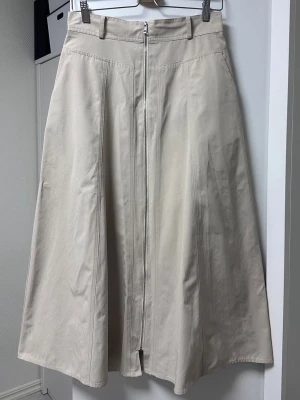 Beige långkjol med dragkedja - Stilren beige långkjol med markerad söm och synlig dragkedja framtill. Kjolen har en A-linjeformad siluett och bälteshällor i midjan. Perfekt för dig som gillar minimalistisk och clean stil.