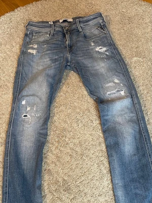 Blå slitna anbass jeans från Replay - Säljer ett par blå skinny jeans från Replay med slitningar och hål på både lår och knän. Jeansen har klassisk femficksmodell, låg midja och detaljerade sömmar. Perfekta för en avslappnad och trendig look.