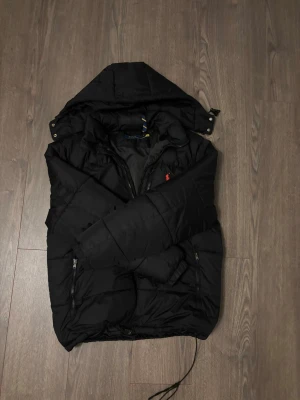 Svart pufferjacka från Polo Ralph Lauren - Säljer en svart pufferjacka från Polo Ralph Lauren med huva och dragkedja framtill. Jackan har två sidofickor med dragkedja och den klassiska röda loggan broderad på bröstet. Perfekt för kalla vinterdagar och riktigt skön att bära.