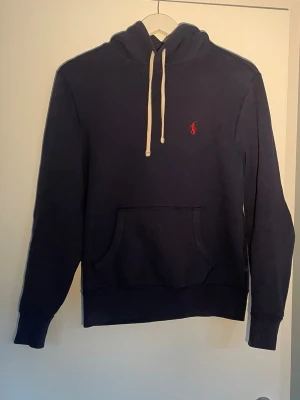 Mörkblå hoodie från Polo Ralph Lauren - Mörkblå hoodie från Polo Ralph Lauren med klassisk känguruficka och vit dragsko i huvan. Ikoniska röda loggan broderad på bröstet. Tillverkad i mjukt bomullsmaterial som är perfekt för en avslappnad stil.