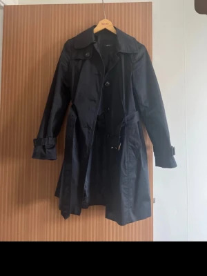 Svart trenchcoat från Jake*s - Stilren svart trenchcoat från Jake*s med klassisk dubbelknäppning, bälte i midjan och justerbara ärmslut. Jackan har bred krage och är tillverkad i ett glansigt tyg som ger en snygg finish. Perfekt för dig som vill ha en tidlös och trendig look.