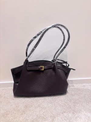 Mörkbrun handväska med gulddetalj - Snygg mörkbrun handväska i slätt läder med guldfärgat spänne framtill och dubbla axelband. Väskan har dragkedja upptill och rymligt fack inuti. Perfekt accessoar för dig som gillar stilrena detaljer och klassisk design.