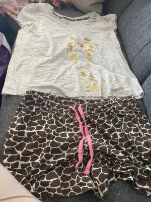 Giraffmönstrad pyjamas med shorts - Pyjamas med ljusgrå t-shirt med guldfärgad girafftryck och liten rosa rosett, samt bruna shorts med vitt giraffmönster och rosa dragsko. Perfekt för chillkvällar och mysiga morgnar. Materialet känns mjukt och bekvämt mot huden. Stl 48/50