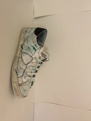  Custom Philippe model skor - Säljer ett par vita Philippe Model sneakers i skinn med gröna färgstänk och svart detalj på hälen. Skorna har klassisk snörning och låg profil. Perfekta för dig som gillar en unik och kreativ look.