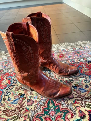 Bruna klassiska cowboy boots - Säljer ett par bruna cowboy boots i äkta läder med dekorativa sömmar längs skaftet och foten. De har spetsig tå och medelhög klack, samt typisk western-design. Perfekta för dig som vill ha en tidlös westernstil.