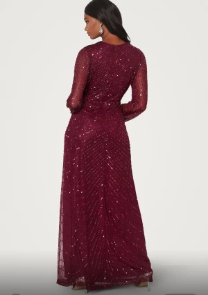 Vinröd långklänning med paljetter - Vinröd långklänning med glittriga paljetter och v-ringning. Klänningen har långa, transparenta ärmar och markerad midja med lätt omlott-effekt framtill. Perfekt för speciella tillfällen när du vill sticka ut.