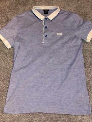 Blå polotopp från Hugo Boss - Säljer en snygg blå polotopp från Hugo Boss med vit krage och vita detaljer vid ärmslut. Toppen har tre blå knappar och logotypen BOSS HUGO BOSS broderad på bröstet. Materialet är mjukt och känns som bomull, perfekt för en stilren look.