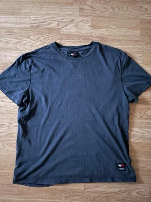 Mörkgrå t-shirt från Tommy Hilfiger - Säljer en mörkgrå t-shirt från Tommy Hilfiger med diskret logga på bröstet och en liten patch med flaggan nere vid kanten. Klassisk rund hals och korta ärmar. Perfekt för dig som gillar stilrena basics med en touch av premium.Helt som ny! Öppen för frågor och prisförslag