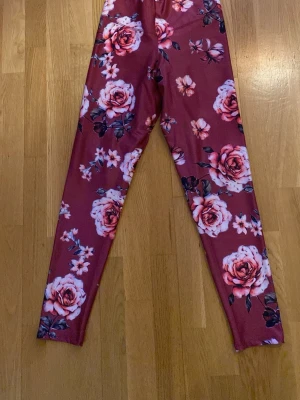 Vinröda leggings med rosor - Snygga vinröda leggings med stort blommönster av rosa och vita rosor samt gröna blad. Tight passform och stretchigt material som sitter skönt. Perfekta för dig som vill sticka ut med ett färgglatt och trendigt plagg.