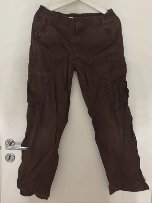 Bruna cargobyxor med många fickor - Säljer ett par bruna cargobyxor med flera stora fickor på sidorna och avslappnad passform. Byxorna har elastisk midja och är tillverkade i ett lätt bomullsmaterial. Perfekta för dig som gillar en chill och praktisk stil.