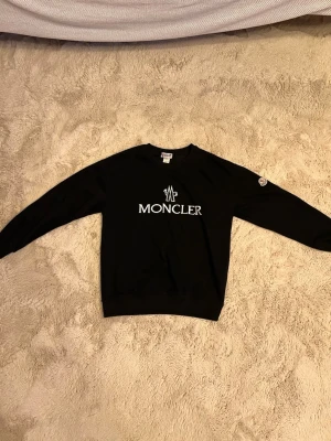 Svart stickad tröja från Moncler - Snygg svart stickad tröja från Moncler med stort vitt Moncler-tryck på bröstet och en liten logga på ärmen. Tröjan har rund halsringning och långa ärmar. Perfekt för dig som gillar stilrena och exklusiva plagg.