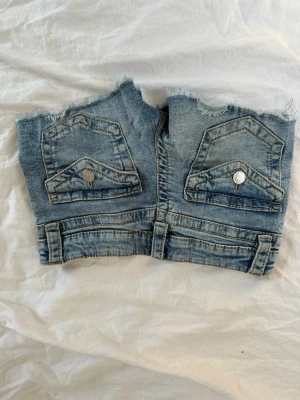 Blå jeansshorts från Ginatricot - Säljer ett par ljusblå jeansshorts från Ginatricot med råa kanter och två bakfickor med lock och silverfärgade knappar. Klassisk denimlook med bälteshällor och slitna detaljer för en avslappnad vibe.