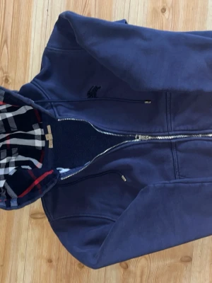 Marinblå hoodie från Lacoste - Snygg marinblå hoodie från Burberry  med dragkedja och logga på bröstet. Huvan är fodrad med rutigt tyg i blått, vitt och rött för en extra cool detalj. Perfekt för dig som gillar stilrena och bekväma plagg. PRISET KAN DU DISKUTERAS VID SNABB KÖP🤙🏽💯
