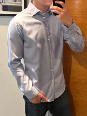 Ljusblå skjorta med knappar - Säljer en stilren ljusblå skjorta med klassisk krage och vita knappar. Skjortan har lång ärm och en normal passform, perfekt till jeans eller chinos. Tillverkad i mjuk bomull för en bekväm känsla hela dagen.
