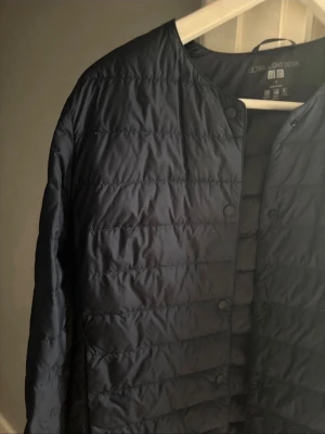 Uniqlo jacka - Snygg mörkblå dunjacka från Uniqlo Ultra Light Down. Jackan är quiltad, har tryckknappar framtill och en minimalistisk design utan krage. Perfekt för lager-på-lager och enkel att bära med sig tack vare den lätta vikten.