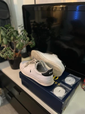 Vita Polo Ralph Lauren sneakers - Snygga vita sneakers från Polo Ralph Lauren med svart hälparti och klassisk logga på sidan. Skorna har lågt skaft och vita snören, och kommer med extra skosnören. Perfekta för en clean och stilren look.