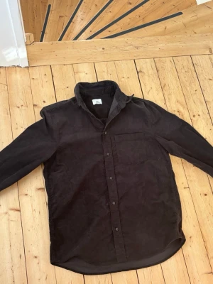 Svart manchesterskjorta från H&M - Säljer en svart manchesterskjorta från H&M med klassisk krage, bröstficka och knappar framtill. Skjortan har lång ärm och är tillverkad i mjuk manchester som ger en schysst vintagekänsla. Perfekt att styla lager på lager.