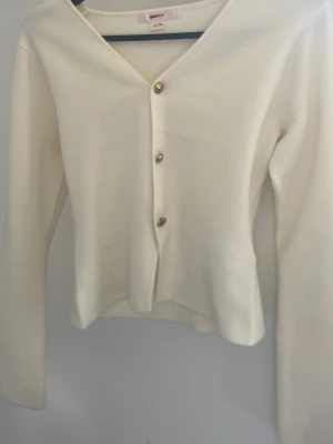 Vit kofta med guldknappar från Bik Bok - Stilren vit kofta från Gina tricot  med v-ringning och tre dekorativa guldknappar framtill. Koftan har lång ärm och är tillverkad i ett mjukt, tunt stickat material som ger en clean och fräsch look. Perfekt att matcha med jeans eller kjol.