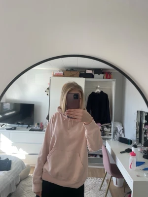 Ljusrosa hoodie med huva - Säljer en ljusrosa hoodie med klassisk känguruficka och dragsko i huvan. Hoodien har en avslappnad passform och ribbade muddar vid ärmar och nederkant. Perfekt för en chill och bekväm stil. Materialet känns mjukt och skönt mot huden.