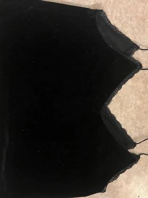 Svart sammetslinnetopp med spets - Snygg svart linnetopp i mjuk sammet med tunna axelband och spetsdetaljer längs v-ringningen. Perfekt att styla lager på lager eller bära som den är för en trendig look.