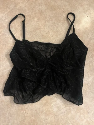 Svart spetsig croppad linnetopp - Snygg svart croppad linnetopp med tunna axelband och genomskinlig spets med blommönster. Perfekt för att lyfta vilken outfit som helst med en edgy touch.