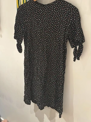 Svart prickig kortklänning Monki - Kort svart klänning från Monki med färgglatt prickigt mönster i vitt, rosa, blått och orange. Klänningen har korta ärmar med knyt i sidan och en rak passform. Perfekt för dig som gillar lekfulla detaljer och enkel stil.