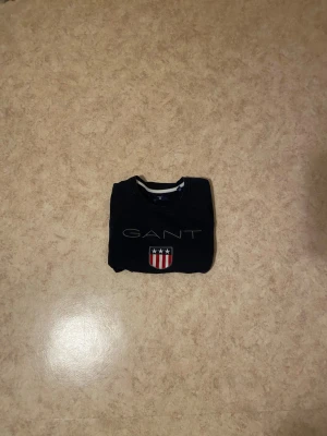 Mörkblå sweatshirt från GANT - Snygg mörkblå sweatshirt från GANT med klassisk logga och ett broderat sköldmärke med stjärnor och ränder på bröstet. Tröjan har rund halsringning och ribbade muddar vid ärmslut och nederkant. Perfekt för en avslappnad och stilren look.